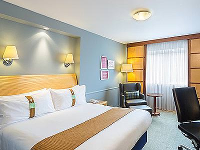 A double bed with a throw on top in a blue hotel room, facing a desk chair, with a chair in the corner