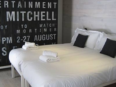 A double bed topped with black cushions and towels, with a black and white wall in the background