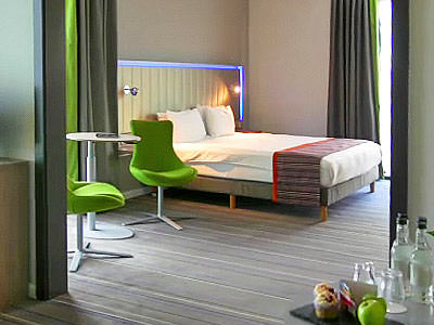 A double bed, with green chairs and a table in the foreground 