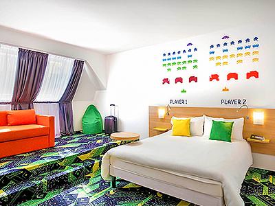 A white double bed with an orange sofa in the background, in a room with a vibrant green carpet and Space Invader stencils on the wall - Rank #5