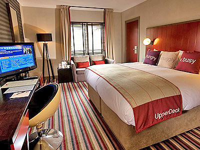 A double bed in a hotel room, topped with a throw and three cushions, facing a turned on TV, desk and desk chair with a grey sofa in the back - Rank #7