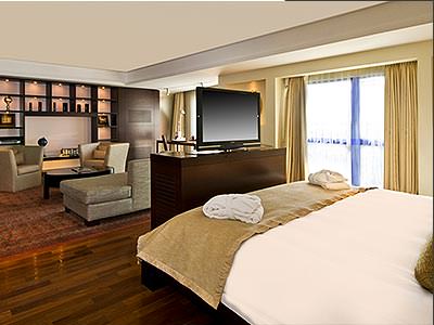 A double bed topped with a throw and towels, facing a TV, with a living area in the background