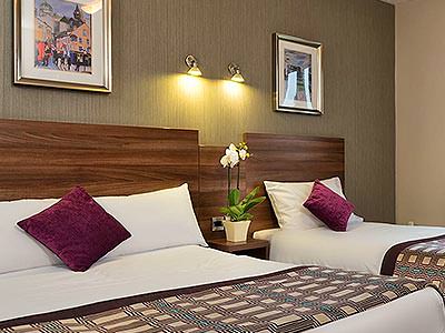 Two double beds in a hotel room, topped with throws and pink cushions, with a bedside table in between both beds