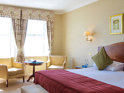 A double bed with a red throw and cushions, with two chairs and a table in front of the window