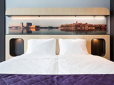Close up of a white double bed, topped with a purple throw, with a pitcure above the bed
