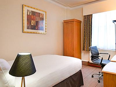 A lamp in the foreground of a double room with a desk chair and a small, thin wardrobe 