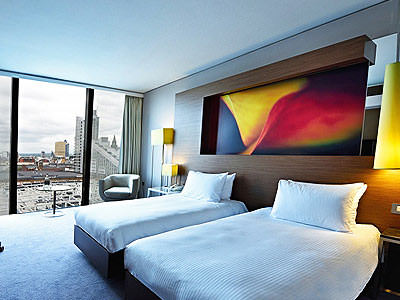 Two white single beds, with a painting above the beds, in a hotel room with floor to ceiling windows 