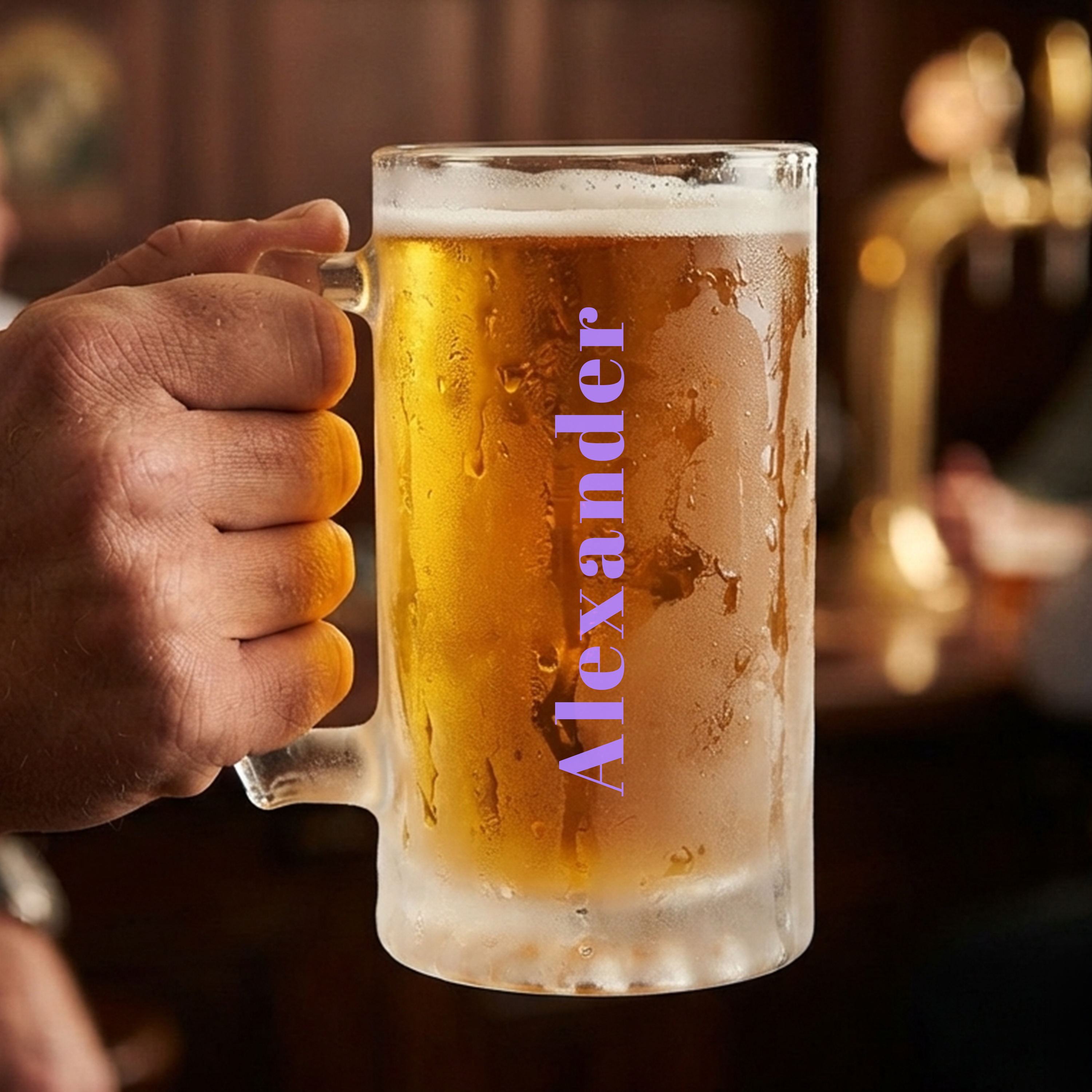 Someone holding a personalised stein in a pub setting with a sturdy glass handle, frosted glass and custom text reading Alexander that runs vertically up the glass