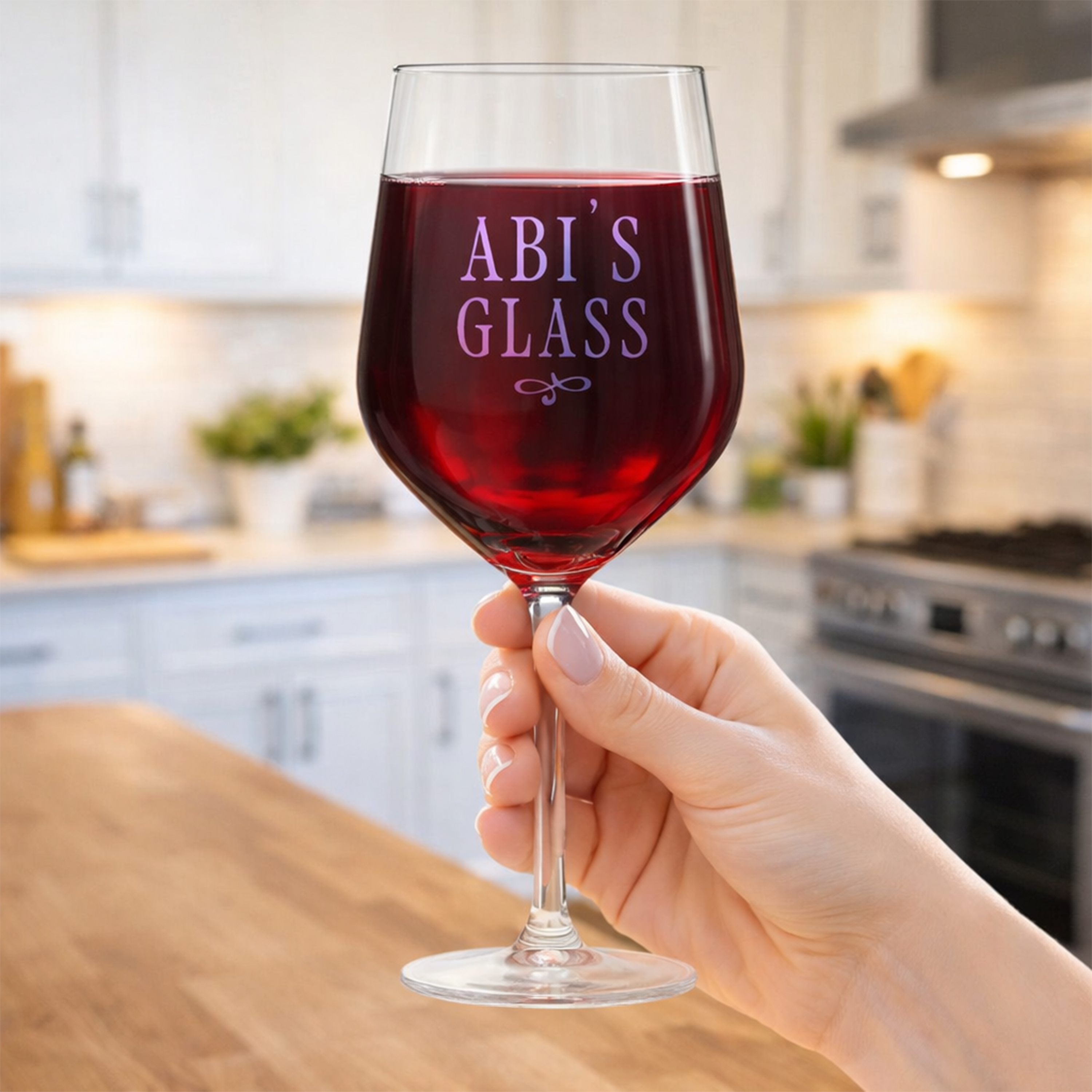 A customisable wine glass full of red wine that has been personalised with the custom text Abi's Glass with a background of a kitchen
