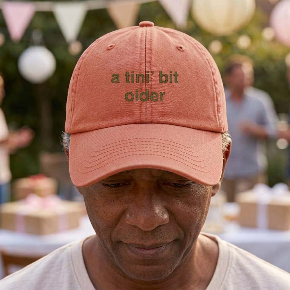 A female model wearing a premium hen do cap in orange that has been embroidered with the phrase a tini's bit older in a green font in front of a blurred garden party background