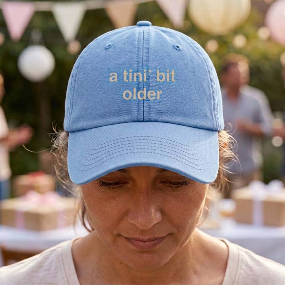 A female model wearing a premium hen do cap in blue that has been embroidered with the phrase a tini's bit older in a tusk font in front of a blurred garden party background