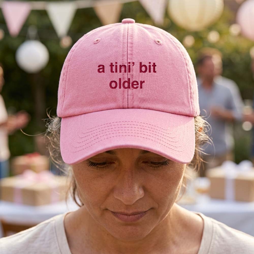 A female model wearing a premium hen do cap in pink that has been embroidered with the phrase a tini's bit older in a red font in front of a blurred garden party background