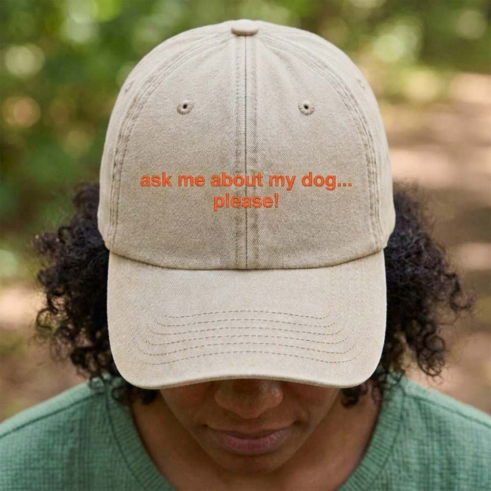 A female model stood in front of an outdoor background wearing a premium cap in sand that has been embroidered with the custom phrase ask me about my dog....please! in an orange font