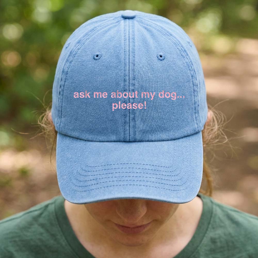 A female model stood in front of an outdoor background wearing a premium cap in light blue that has been embroidered with the custom phrase ask me about my dog....please! in a pink font