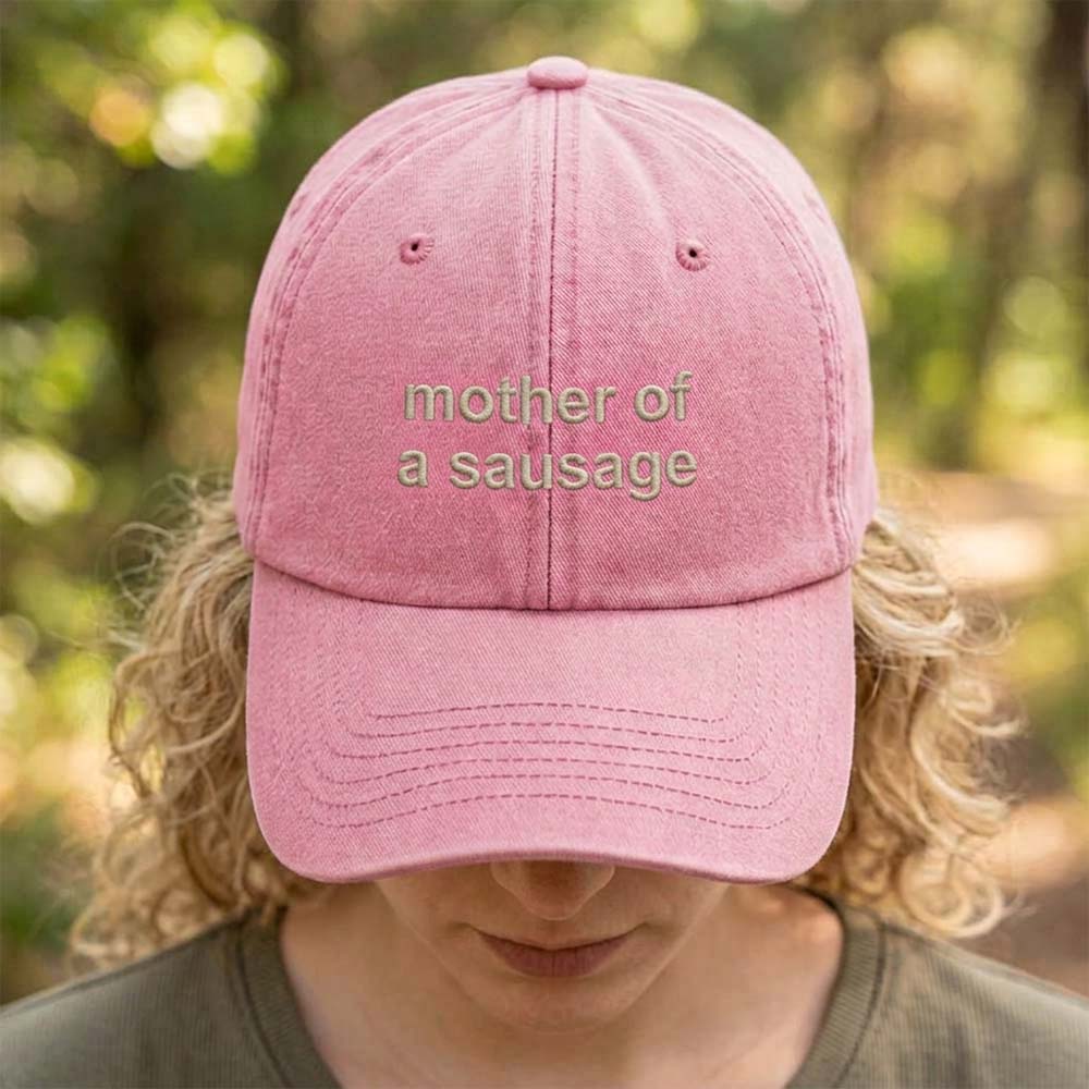 A female model stood in front of a blurred forest background wearing a premium vintage cap in pink that has been embroidered with the custom phrase mother of a sausage in a tusk font