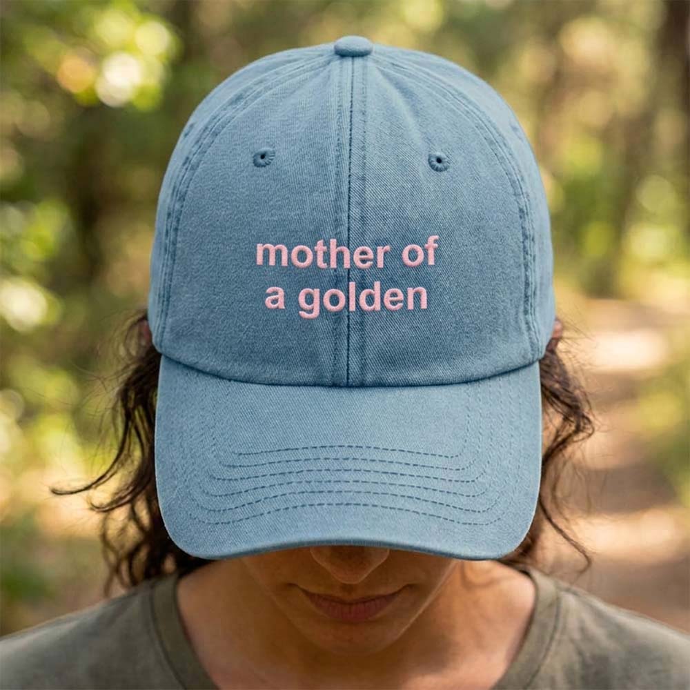 A female model stood in front of a blurred forest background wearing a premium vintage cap in blue that has been embroidered with the custom phrase mother of a golden in a pink font