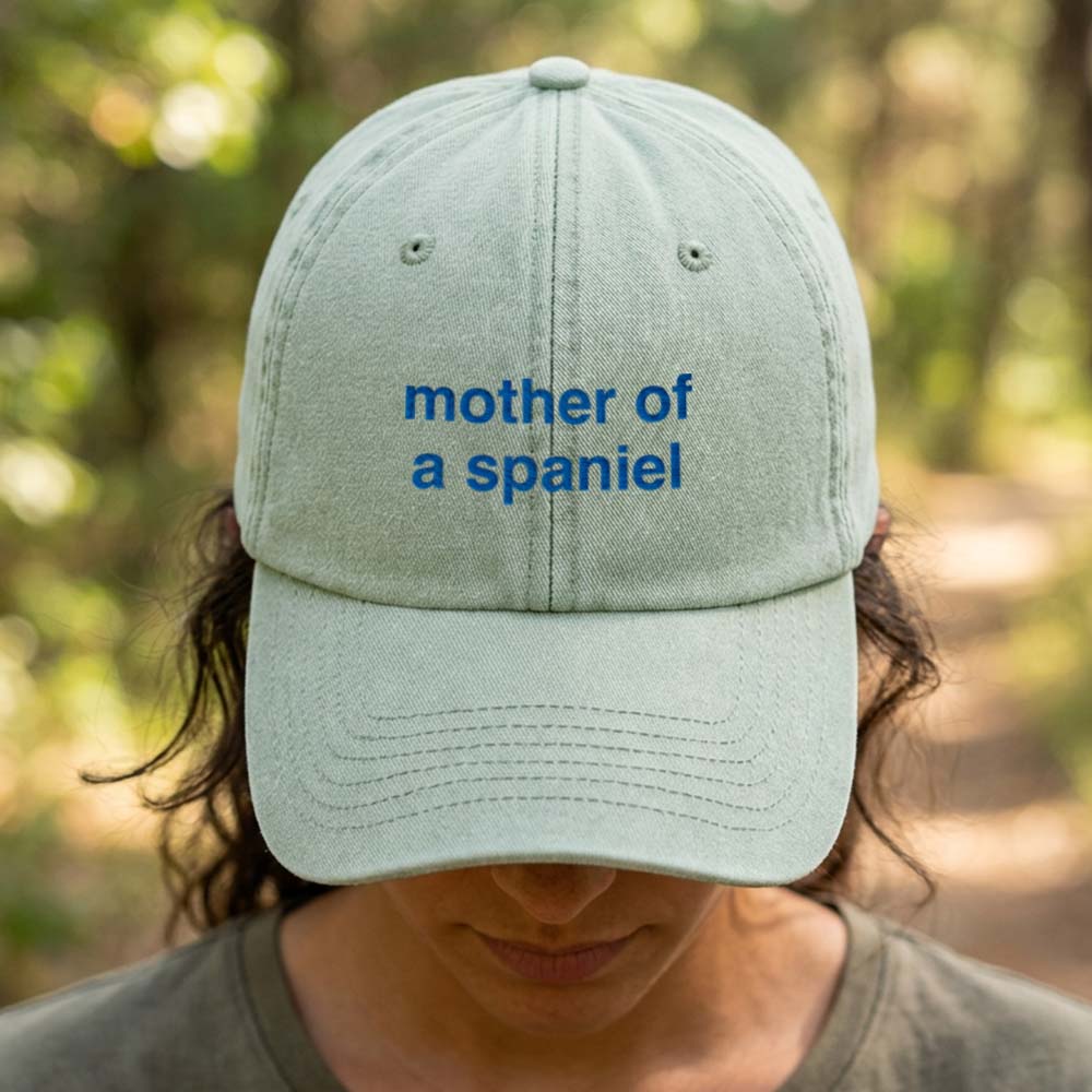 A female model stood in front of a blurred forest background wearing a premium vintage cap in green that has been embroidered with the custom phrase mother of a spaniel in a blue font