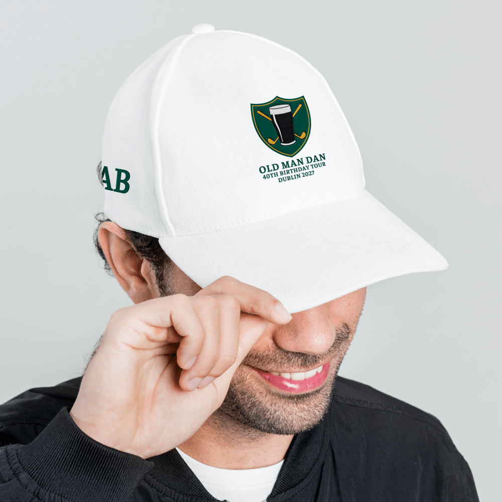 A side view of male model wearing a white golfing cap with an embroidered crest that has a pint of Guinness in front of golf clubs that cross over each other in an X shape with three lines of custom text that read Old Man Dan, 40th Birthday Tour, Dublin 2027 as well as the initials AB embroidered on the side