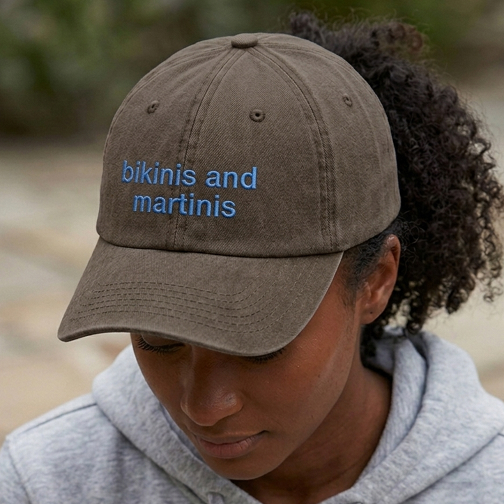 A female model wearing a premium hen do cap in brown that has been embroidered with the phrase bikinis and martinis in a blue font