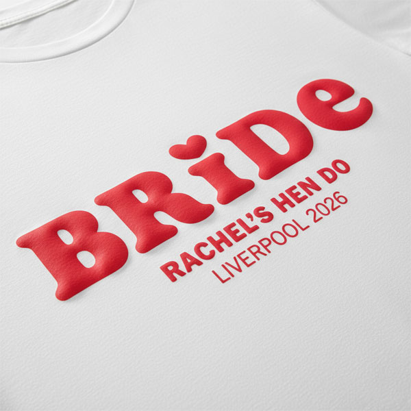 A white hen do shirt with custom pink 'puff print' bubble writing that reads Bride (with a heart above the I) as well as Rachel's Hen Do, Liverpool 2026