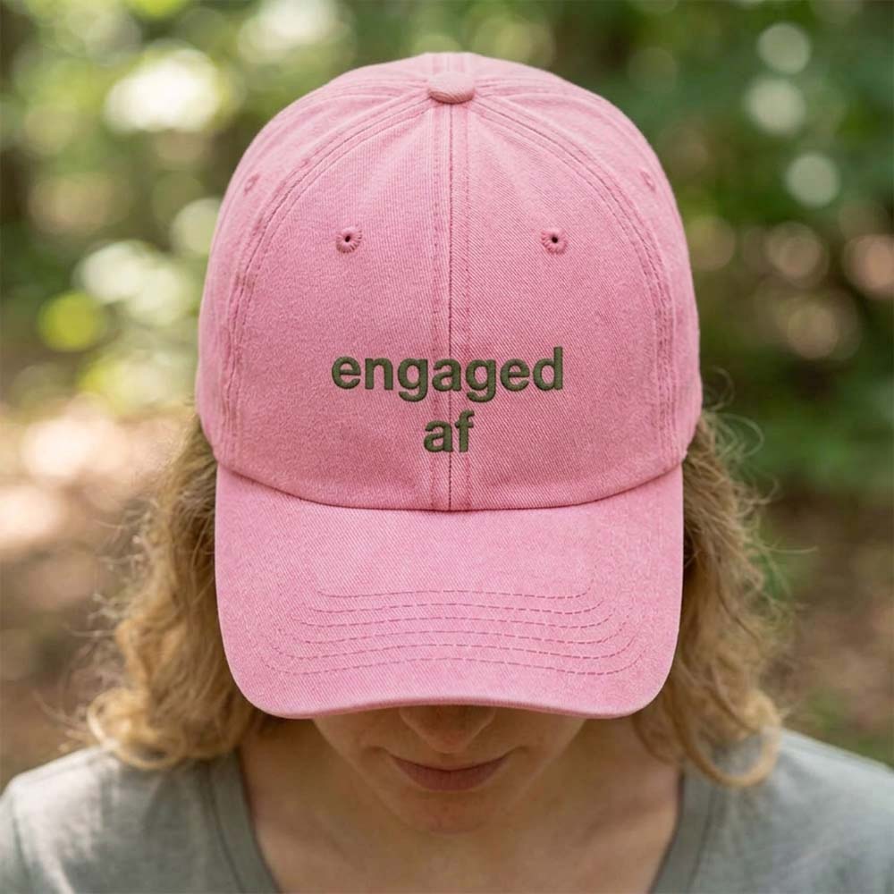 A female model wearing a premium hen do cap in pink that has been embroidered with the phrase engaged af in a green font with a background of a blurred out forest