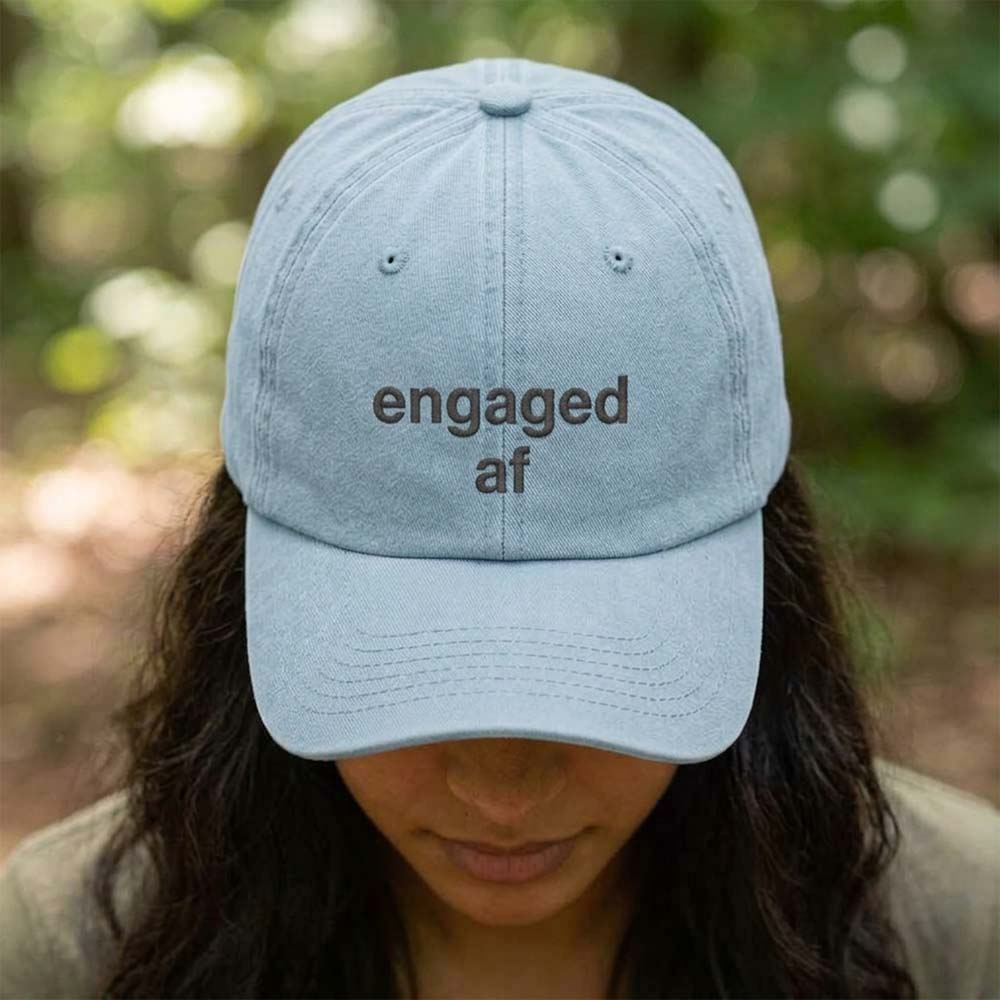 A female model wearing a premium hen do cap in light blue that has been embroidered with the phrase engaged af in a navy font with a background of a blurred out forest