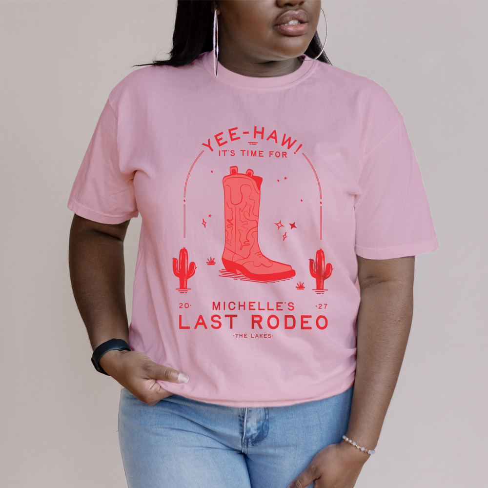 A burgundy t-shirt with a pink design that has a cowboy boot on it and two cactus that reads yee-haw it's time for Megan's last rodeo. 