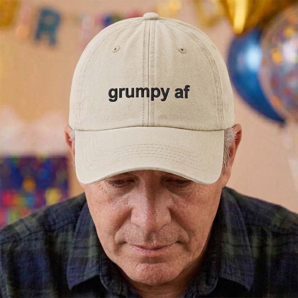 A male model stood in front of a blurred birthday background and wearing a premium birthday cap in sand that has been embroidered with the phrase grumpy af in a black font