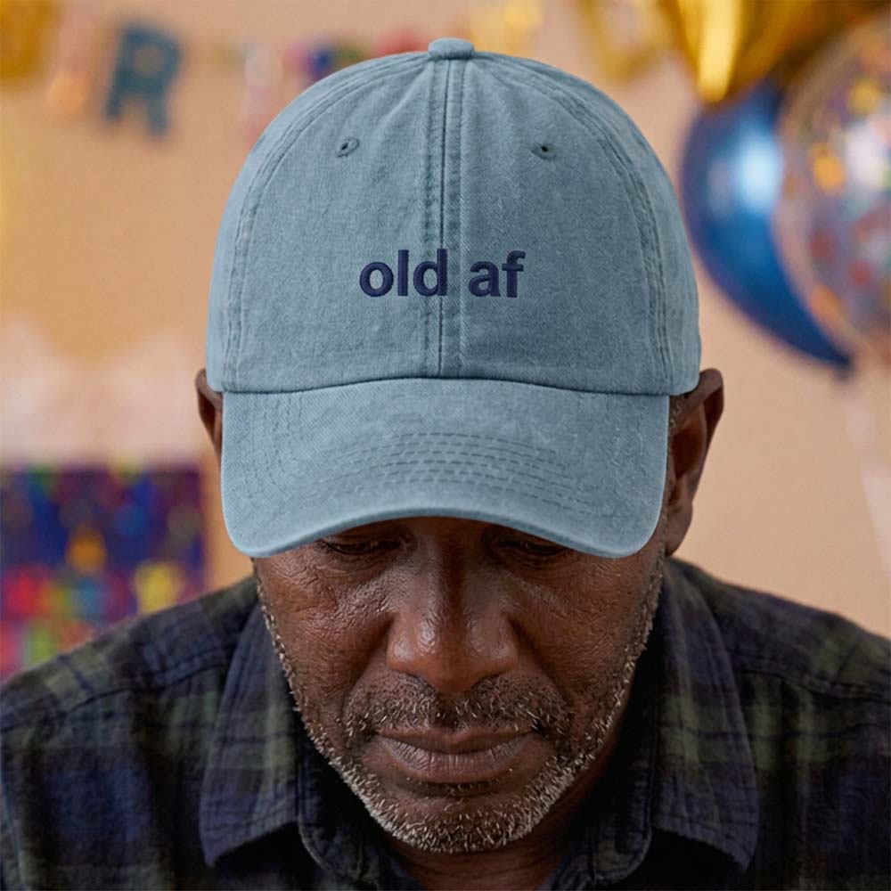 A male model stood in front of a blurred birthday background and wearing a premium birthday cap in blue that has been embroidered with the phrase old af in a navy font