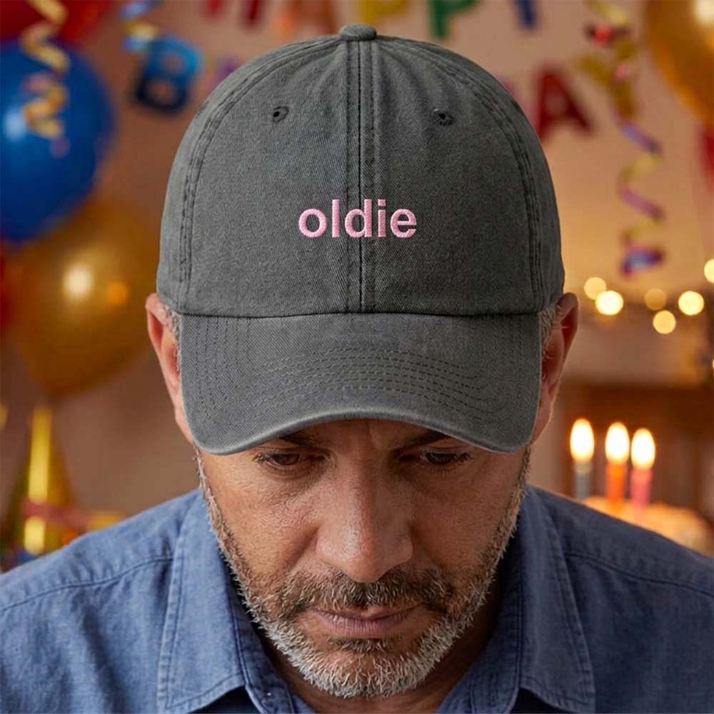 A male model stood in front of a blurred birthday background wearing a premium birthday cap in brown that has been embroidered with the phrase oldie in a pink font