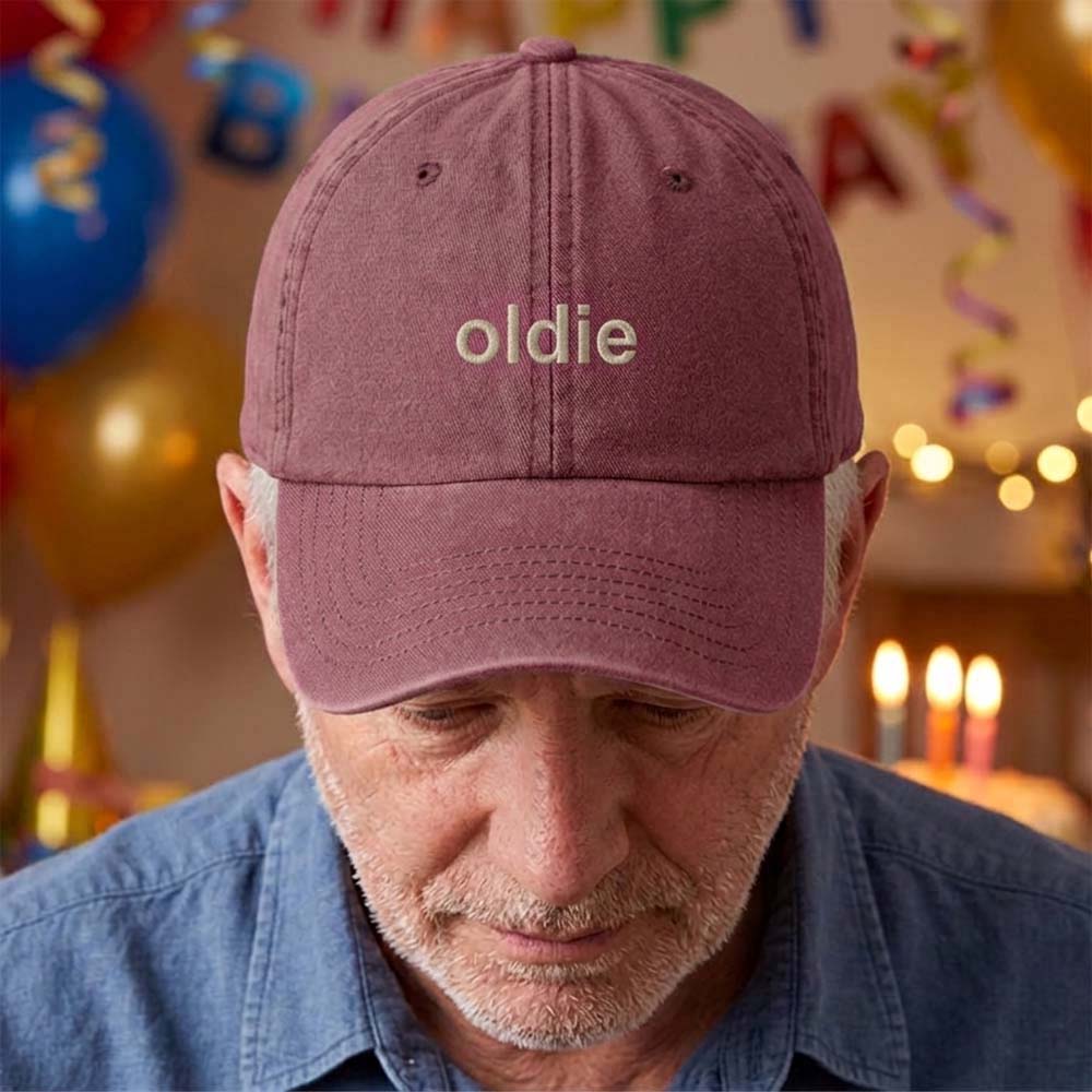 A male model stood in front of a blurred birthday background wearing a premium birthday cap in red that has been embroidered with the phrase oldie in a tusk font