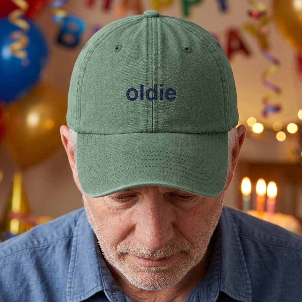 A male model stood in front of a blurred birthday background wearing a premium birthday cap in green that has been embroidered with the phrase oldie in a blue font