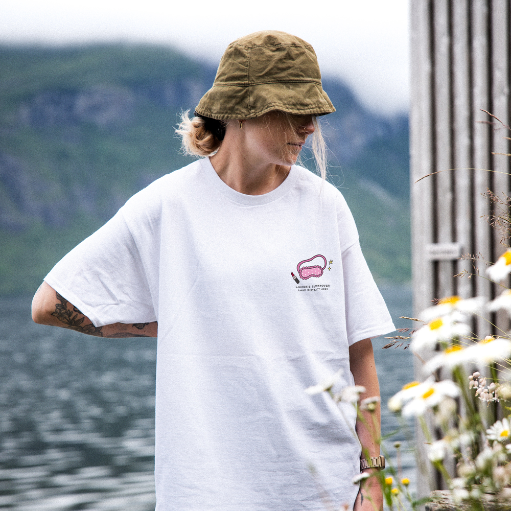 A female model wearing a custom hen do tee that has a sleeping mask and a lipstick drawing on it with the custom text Louise's Sleepover Lake District 2028