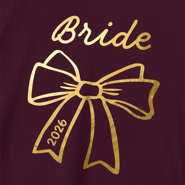 A maroon hen do shirt with a shiny gold foil bow which has the custom text bride above it and 2026 in the bow 