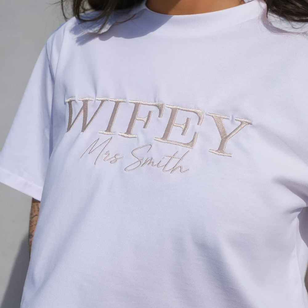 A close up shot of a model wearing a luxurious honeymoon t shirt that has WIFEY embroidered on it in statement text and Mrs Smith embroidered on it in cursive text