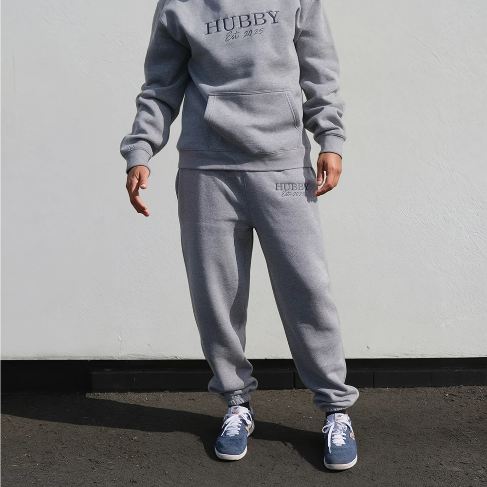 A male model wearing a luxe athletic heather coloured honeymoon hoodie and joggers that both have HUBBY est 2025 embroidered on them 