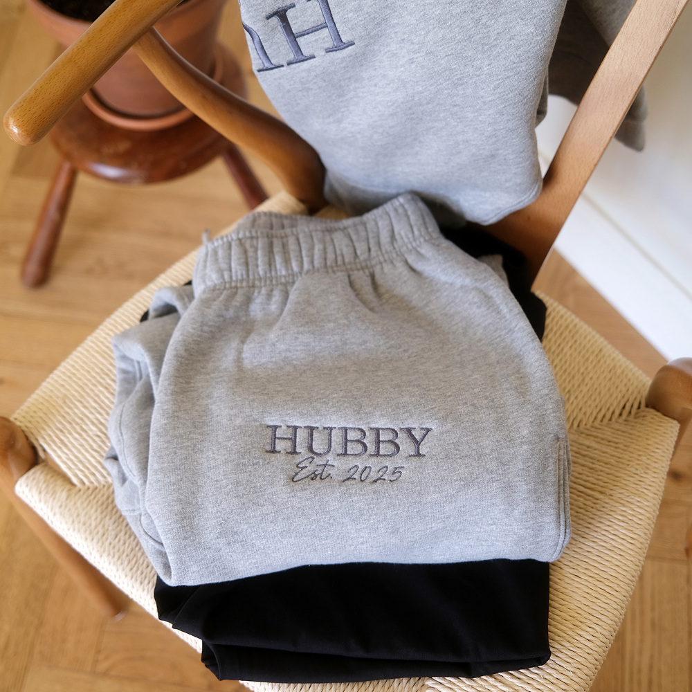 A pair of luxe athletic heather coloured joggers, with embroidered text that reads HUBBY est 2025, that are folded up neatly and sitting on a chair