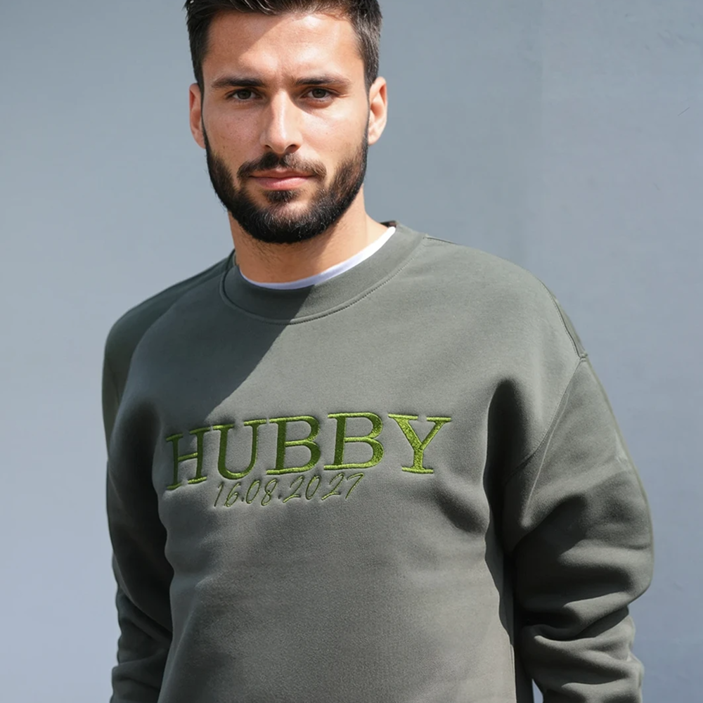 A man wearing a cypress coloured groom honeymoon sweatshirt that has HUBBY and 16.08.2027 underneath it in custom embroidered text 