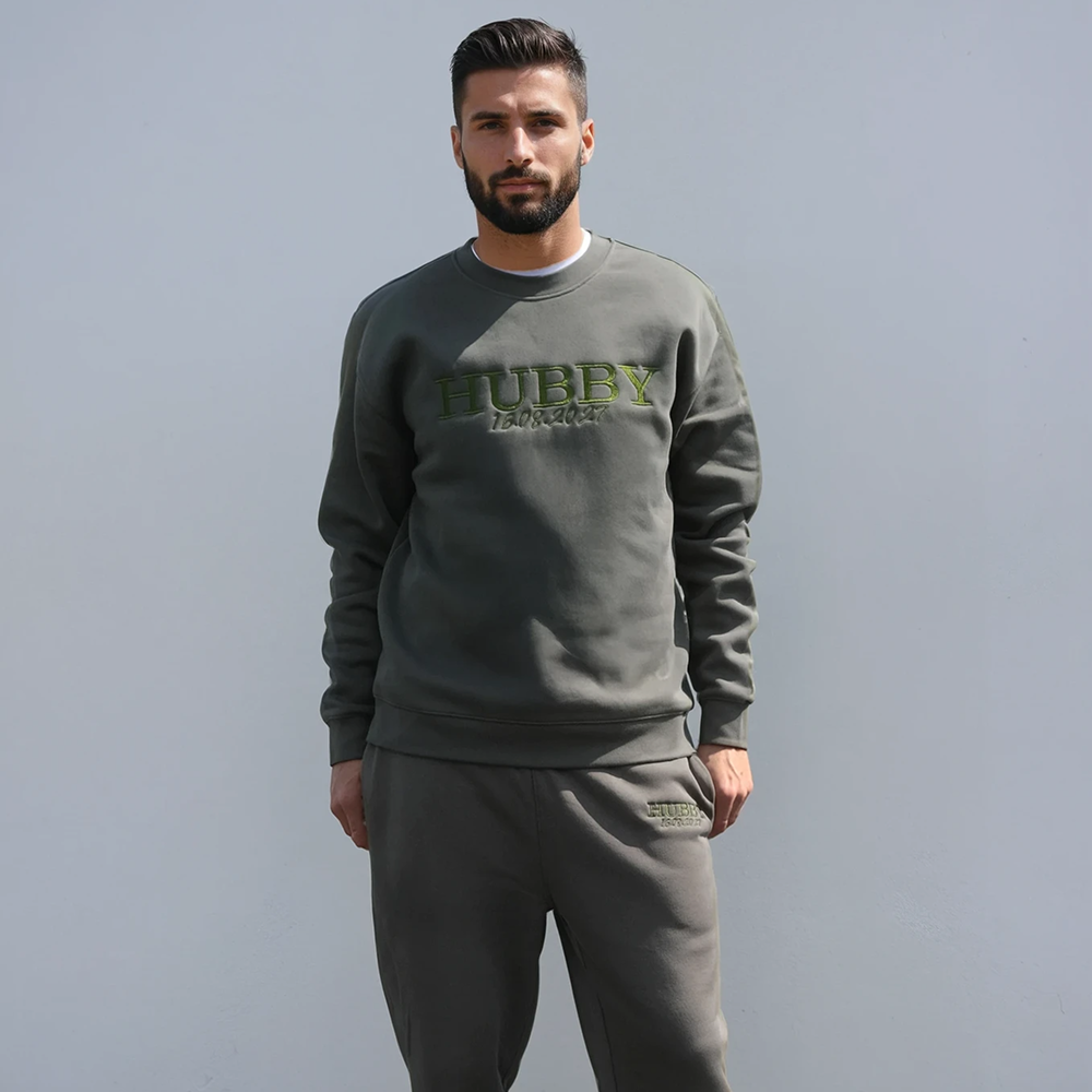 A man wearing a cypress coloured groom honeymoon sweatshirt that has HUBBY and 16.08.2027 underneath it in custom embroidered text 
