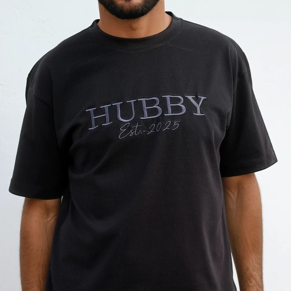 A male model wearing a luxe black honeymoon t shirt with embroidered text that reads HUBBY with Mr Smith underneath it