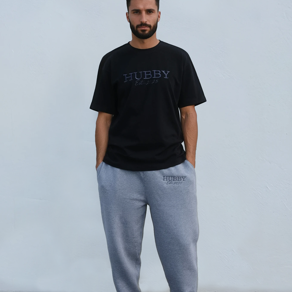 A male model wearing a luxe black honeymoon t shirt with embroidered text that reads HUBBY with Mr Smith underneath it with matching joggers that are an athletic heather colour
