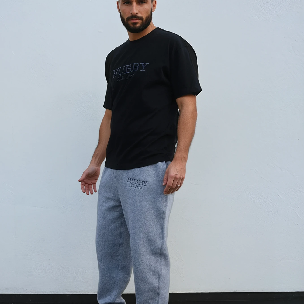 A male model stood sideways wearing a luxe black honeymoon t shirt with embroidered text that reads HUBBY with Est 2025 underneath it with matching joggers that are an athletic heather colour