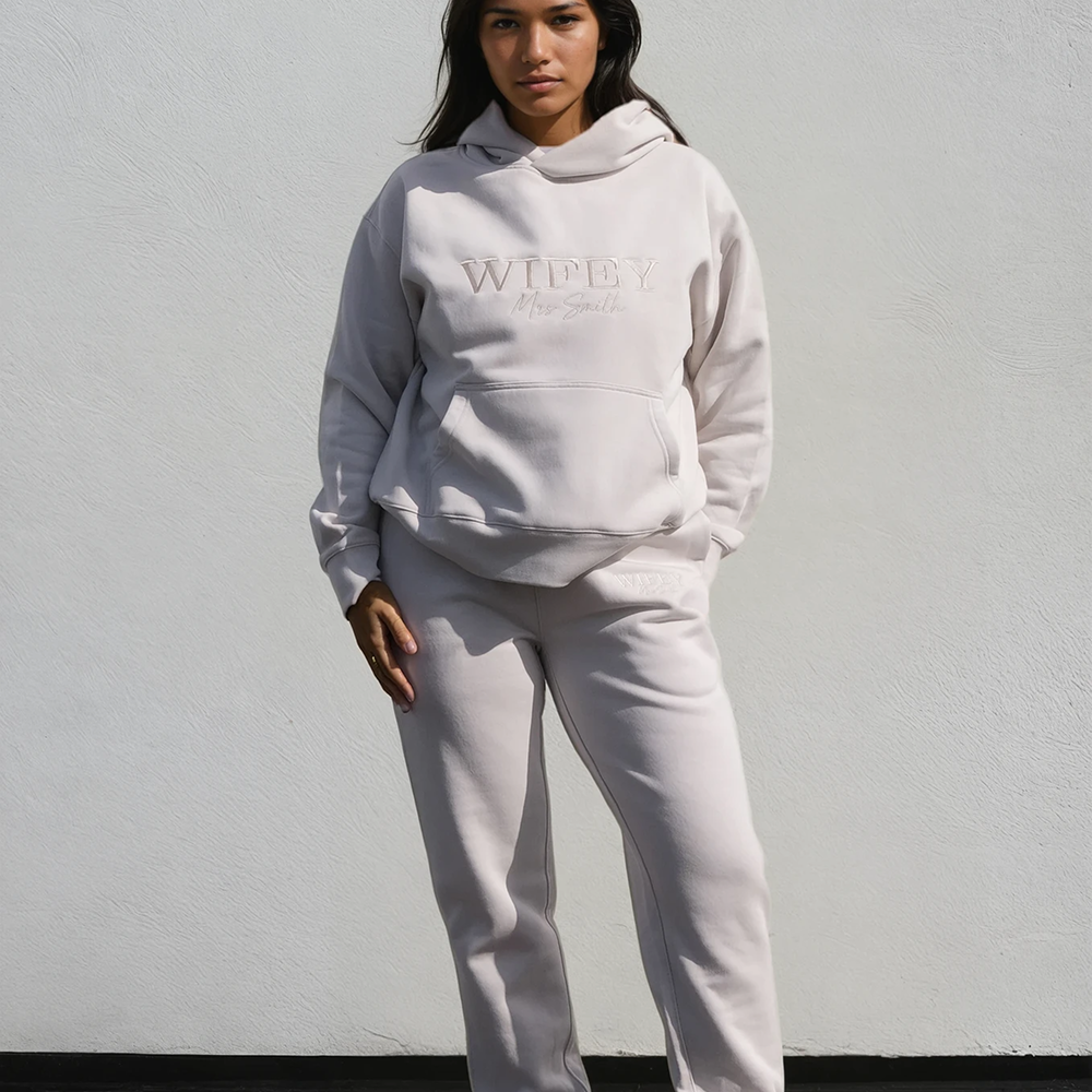 A woman modelling a luxurious looking hoodie that reads WIFEY in statement text and Mrs Smith in cursive with matching joggers