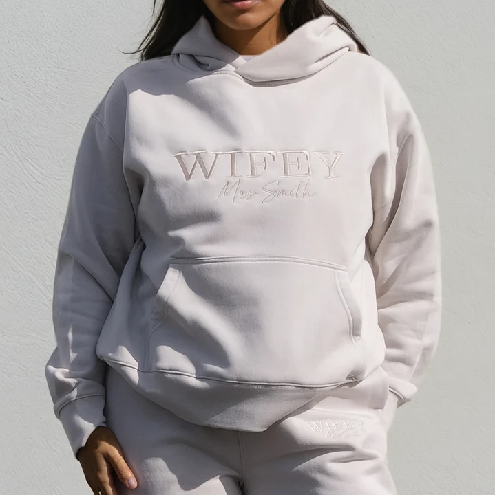 Modelled photo of young woman wearing bone colour honeymoon hoodie with 'WIFEY' and 'Mrs Smith' embroidered on front. Also shows matching joggers.