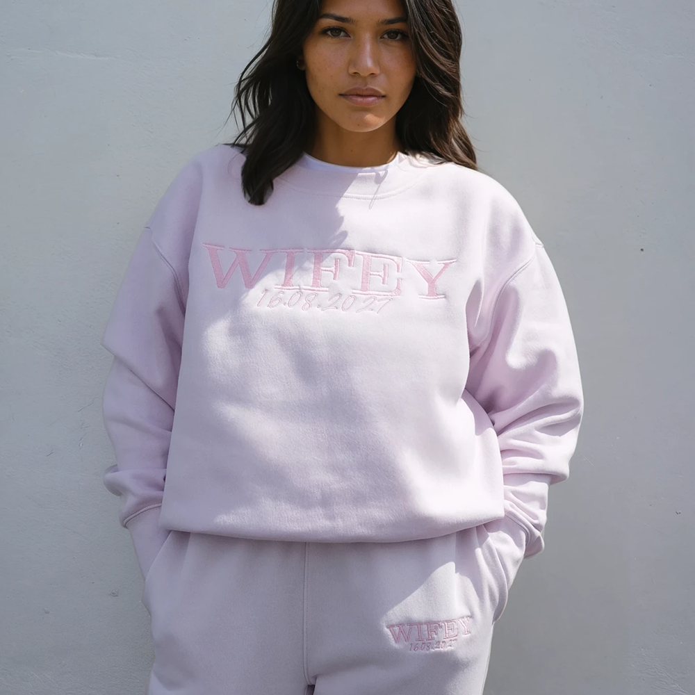  A woman wearing an orchid coloured honeymoon sweatshirt that has WIFEY embroidered on it in a statement font and 16.08.2027 underneath 