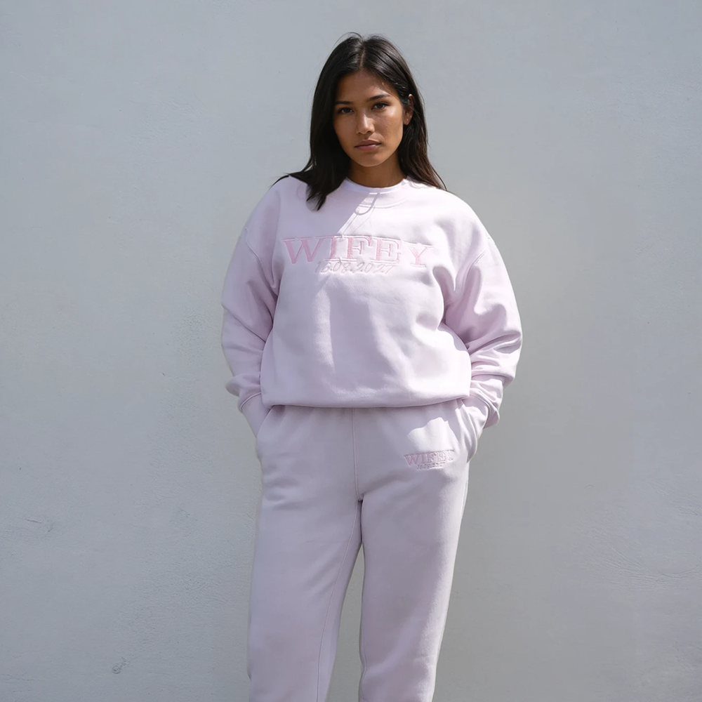 A woman wearing an orchid coloured honeymoon sweatshirt that has WIFEY embroidered on it in a statement font and 16.08.2027 underneath