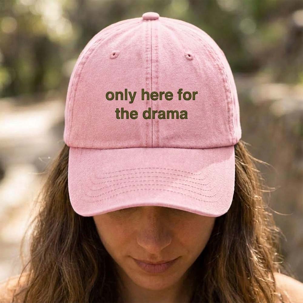 A female model wearing a premium hen do cap in pink that has been embroidered with the phrase only here for the drama in a green font
