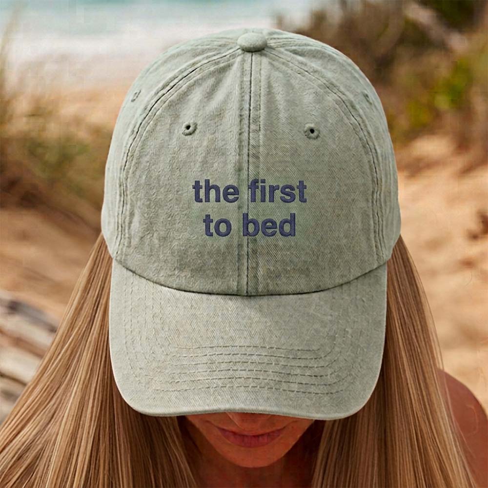 A female model in front of an outdoor background wearing a premium hen do cap in green that has been embroidered with custom text the first to bed in a dark blue font