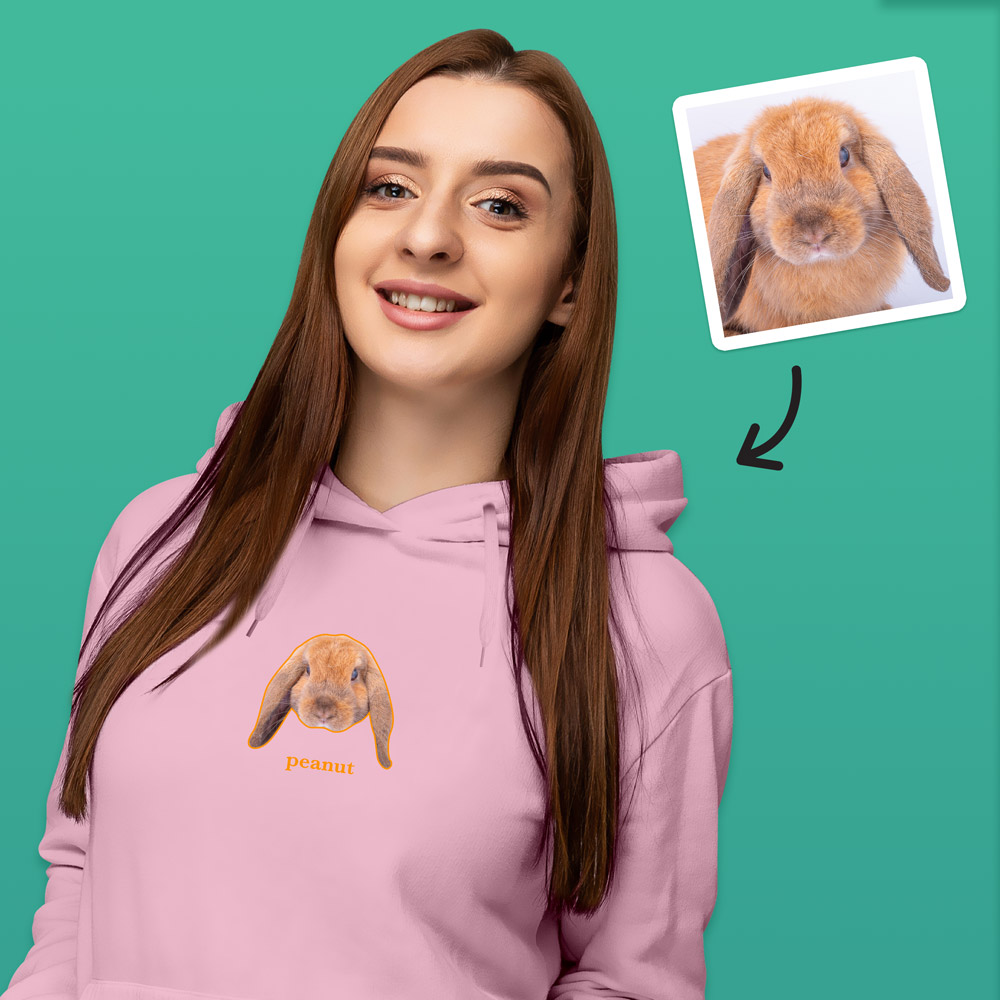A female model wearing a light pink hoodie that can be customised with the face of a pet and up to two lines of custom text which has a rabbit's face on it with the word peanut under it