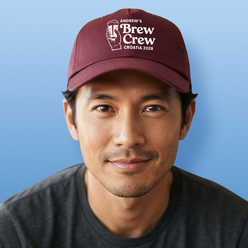 A male model wearing a burgundy custom stag do cap with a graphic of a hand holding a beer and custom text reading Andrew's Brew Crew Croatia 2028 in white font